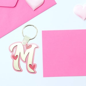 May include: A pink and gold letter "M" keychain with heart accents. The keychain has a silver ring and a leather tab. The background includes pink hearts, a pink envelope, and a pink sticky note on a white surface.