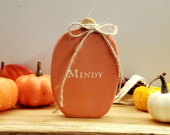 May include: A small, orange pumpkin-shaped wooden decoration with a twine bow and the name "MINDY" written in white paint.