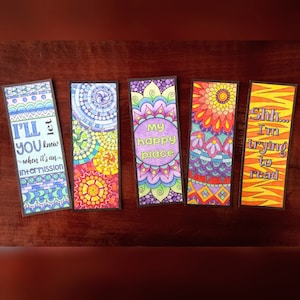 Può includere: Cinque segnalibri colorati con intricati disegni di mandala e citazioni ispiratrici. I segnalibri presentano frasi come "I'll you know when it's an intermission", "My happy place" e "Shh... I'm trying to read".