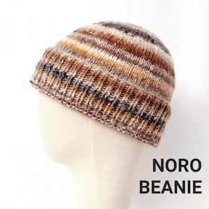 Puede incluir: Un gorro de punto de rayas marrones, beige y grises. El gorro está en una cabeza de maniquí blanca. El texto "Noro Beanie" está impreso debajo del gorro.
