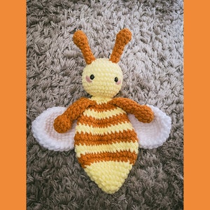 Puede incluir: Un juguete de abeja de crochet con rayas amarillas y marrones, alas blancas y ojos negros. La abeja está tumbada sobre una superficie gris y peluda.