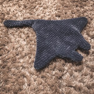 Puede incluir: Un juguete de manta raya de peluche azul oscuro con una superficie texturizada. El juguete está acostado sobre una alfombra de pelo largo marrón claro.