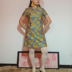 Puede incluir: Vestido de manga corta con un estampado geométrico en amarillo, gris y verde. El vestido se combina con medias blancas y zapatos color crema. La persona lleva pendientes amarillos.