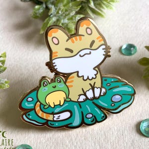 Op de afbeelding: Een goudkleurige emaille pin met een cartoon kat en kikker. De kat is bruin met oranje strepen en witte accenten, zittend op een turquoise waterlelieblad met een groene kikker. De pin heeft een glanzende afwerking en de handtekening.
