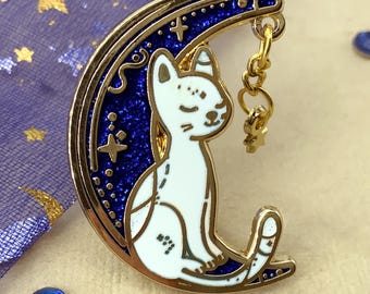 Puede incluir: Un pin de metal dorado con un gato blanco sentado en una luna creciente. La luna tiene un fondo azul brillante con estrellas doradas. Un pequeño dije dorado cuelga de la luna. Un accesorio de temática celestial.