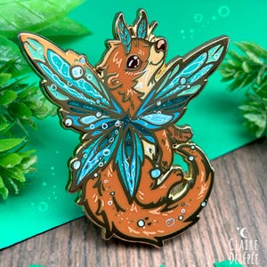 May include: An enamel pin depicting a fantastical creature with orange fur and teal butterfly wings. The pin has gold detailing and is set against a green and brown backdrop. The artist's signature is visible in the bottom right corner.