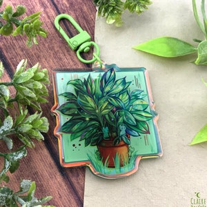 Könnte beinhalten: Ein grüner und weißer Acryl-Schlüsselanhänger mit einer Topfpflanze, in deren Blättern sich kleine weiße Katzen verstecken. Der Schlüsselanhänger hat einen grünen Metallverschluss.