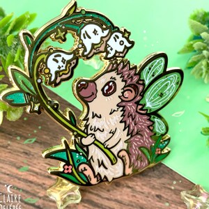 Pode incluir: Um alfinete de esmalte caprichoso com um ouri&ccedil;o com asas de fada, segurando um raminho de flores de l&iacute;rio-do-vale. O alfinete tem um contorno dourado, com detalhes em branco, verde e castanho. O fundo &eacute; verde e de madeira.
