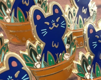 May include: A blue cat enamel pin with a white face and pink ears, sitting in a gold-rimmed orange pot with green leaves and white flowers. The pin is part of a set of similar pins.
