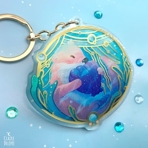 Peut inclure: Un porte-clés en résine transparente représentant une loutre de mer rose et bleue tenant une coquille de mer violette. La loutre est entourée de détails dorés et bleus, notamment des algues et des bulles. Le porte-clés est attaché à un anneau en métal doré.