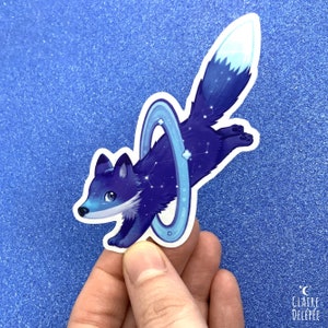 Pode incluir: Um adesivo de raposa azul e branco com um design de galáxia e um anel ao redor. A raposa está pulando através do anel.