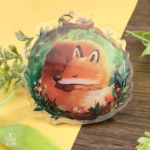 Könnte beinhalten: Ein runder, transparenter Acryl-Anhänger mit einem schlafenden Fuchs, umgeben von grünem Laub, Pilzen und einem kleinen Vogel, der auf einem Ast sitzt. Der Fuchs ist orange und braun mit einer weißen Brust und einem eingerollten Schwanz.