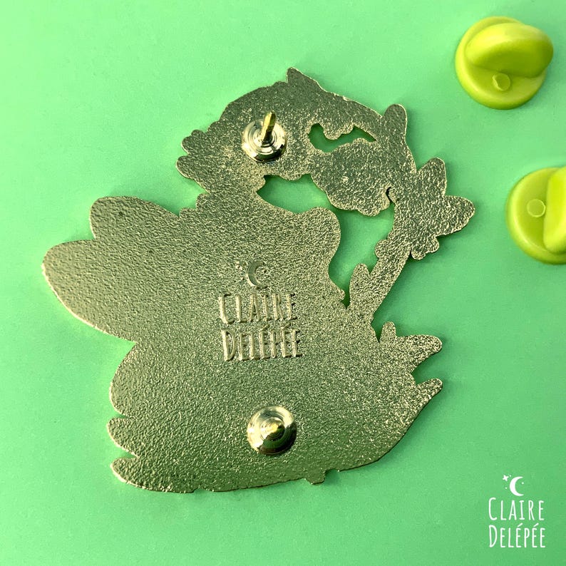 Pode incluir: Parte de tr&aacute;s de um alfinete de esmalte dourado com um design floral. O alfinete tem formato de borboleta com um design floral. O alfinete tem um pino e uma embreagem. O alfinete est&aacute; em um fundo verde. O texto "CLAIRE DEL&Eacute;P&Eacute;E" &eacute; vis&iacute;vel.