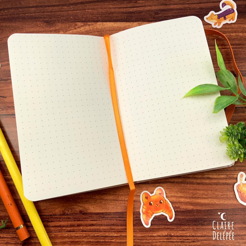 Puede incluir: Un cuaderno blanco con puntos abierto con un marcador de cinta naranja. El cuaderno est&aacute; abierto sobre una superficie de madera con algunas pegatinas de colores en la portada.
