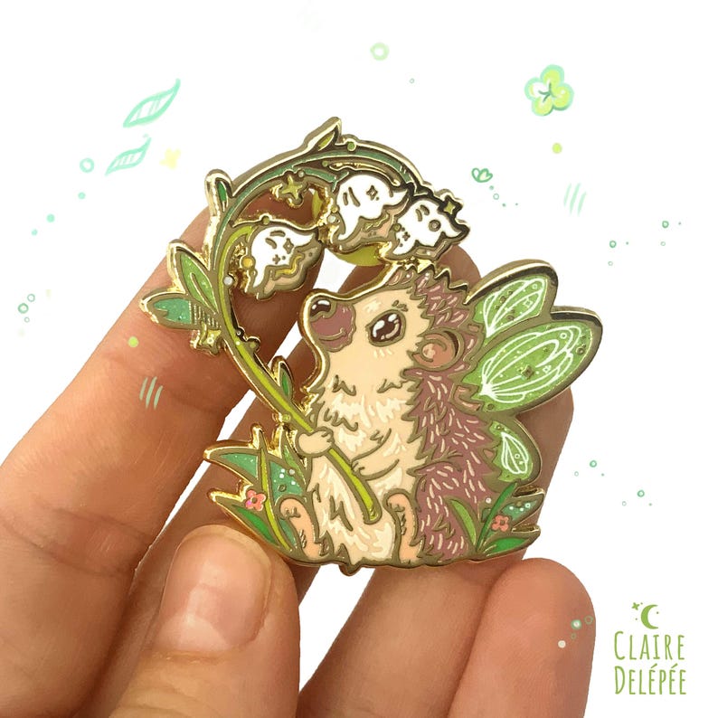Pode incluir: Um pin de esmalte caprichoso com um ouri&ccedil;o com asas de fada, segurando um ramo de l&iacute;rios-do-vale. O pin tem um contorno dourado com detalhes verdes e brancos. O design inclui grama verde e pequenas flores cor-de-rosa.