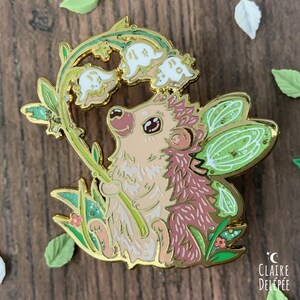 Pode incluir: Um alfinete de esmalte caprichoso com um ouri&ccedil;o com asas de fada, segurando um caule de l&iacute;rio-do-vale. O alfinete tem um contorno dourado com detalhes em verde, rosa e branco. O alfinete est&aacute; sobre um fundo de madeira com folhas decorativas.