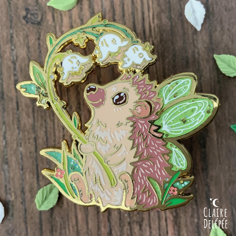Pode incluir: Um alfinete de esmalte caprichoso com um ouri&ccedil;o com asas de fada, segurando um caule de l&iacute;rio-do-vale. O alfinete tem um contorno dourado com detalhes em verde, rosa e branco. O alfinete est&aacute; sobre um fundo de madeira com folhas decorativas.