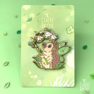 Pode incluir: Pin de esmalte com um ouri&ccedil;o caprichoso com asas de fada, segurando flores de l&iacute;rio-do-vale. O pin tem um contorno dourado, detalhes em rosa e verde e o texto "Claire Delep&eacute;e". O pin est&aacute; em um cart&atilde;o verde.