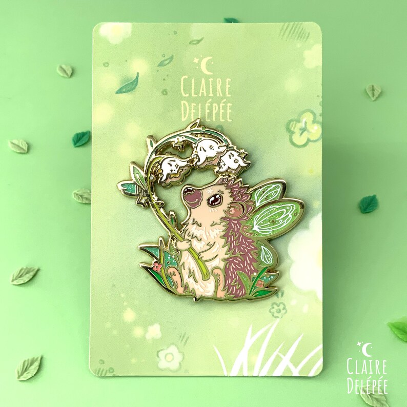 Pode incluir: Pin de esmalte com um ouri&ccedil;o caprichoso com asas de fada, segurando flores de l&iacute;rio-do-vale. O pin tem um contorno dourado, detalhes em rosa e verde e o texto "Claire Delep&eacute;e". O pin est&aacute; em um cart&atilde;o verde.