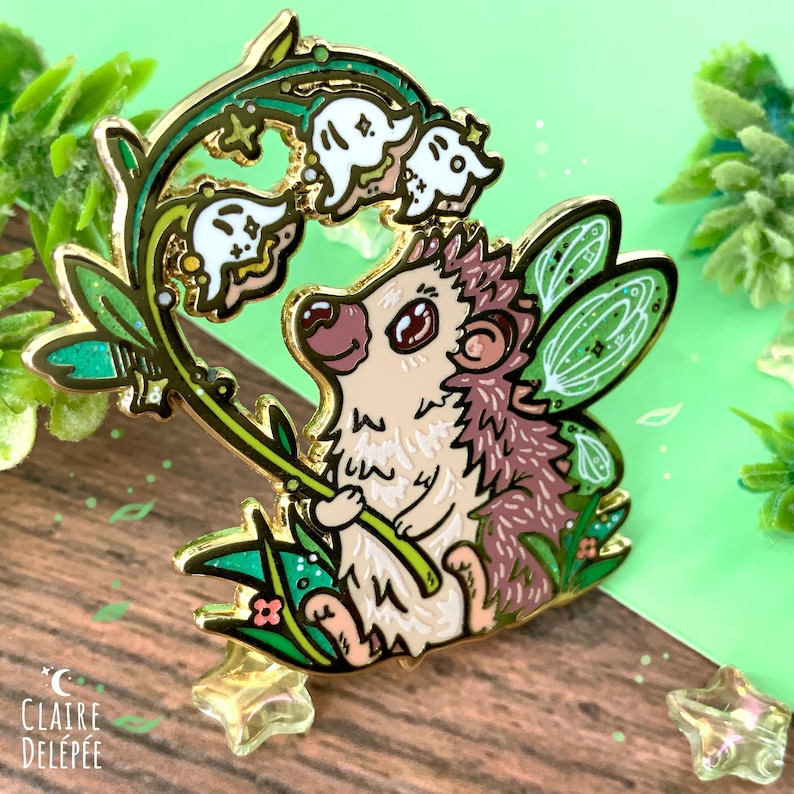 Pode incluir: Um alfinete de esmalte caprichoso com um ouri&ccedil;o com asas de fada, segurando um raminho de flores de l&iacute;rio-do-vale. O alfinete tem um contorno dourado, com detalhes em branco, verde e castanho. O fundo &eacute; verde e de madeira.
