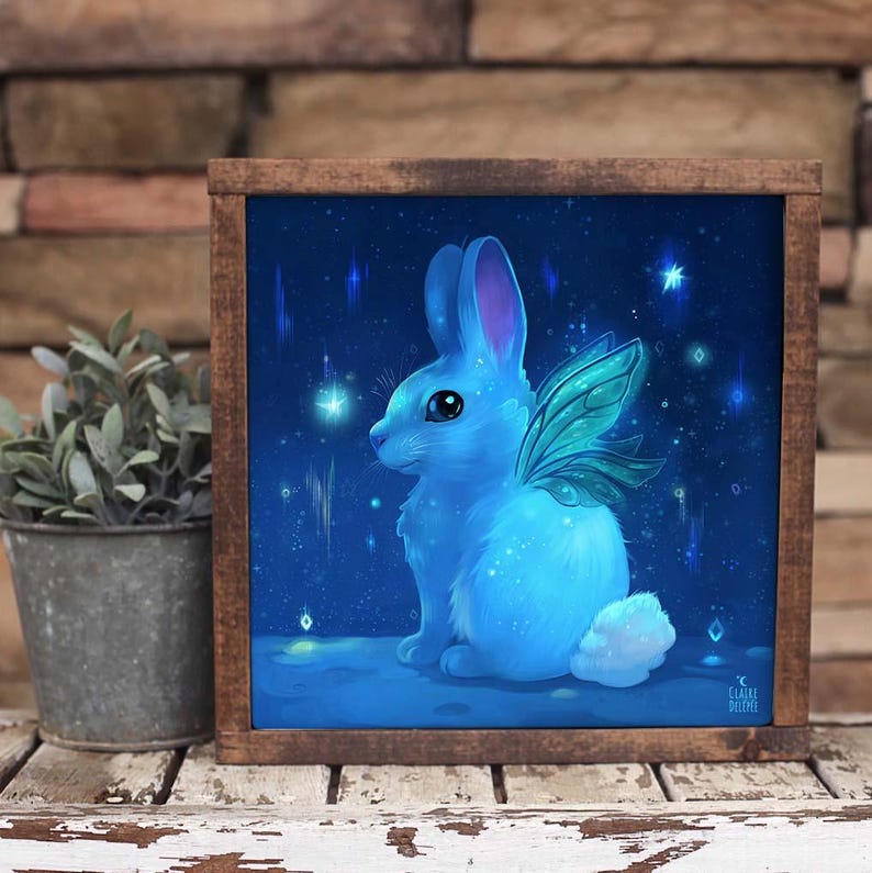 May include: A blue rabbit with fairy wings sits on a blue background with white stars. The rabbit has a light blue body and pink ears. The wings are green and translucent. The background is a dark blue with white stars and a few white streaks.