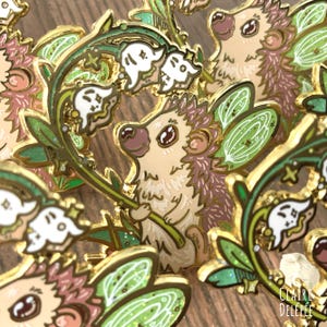 Pode incluir: Pins de esmalte com um design de ouri&ccedil;o caprichoso. Os pins t&ecirc;m um contorno dourado, com o ouri&ccedil;o em tons de bege e castanho, segurando um caule verde com flores brancas. Os pins s&atilde;o decorados com folhas verdes e flores brancas.