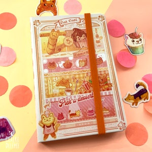 Puede incluir: Un cuaderno ilustrado rosa y naranja con tem&aacute;tica de cafeter&iacute;a de gatos. La portada presenta un gato con un gorro de chef y sosteniendo un batidor, con el texto "Cat Cafe" encima. Hay otras ilustraciones de gatos y postres en la portada.