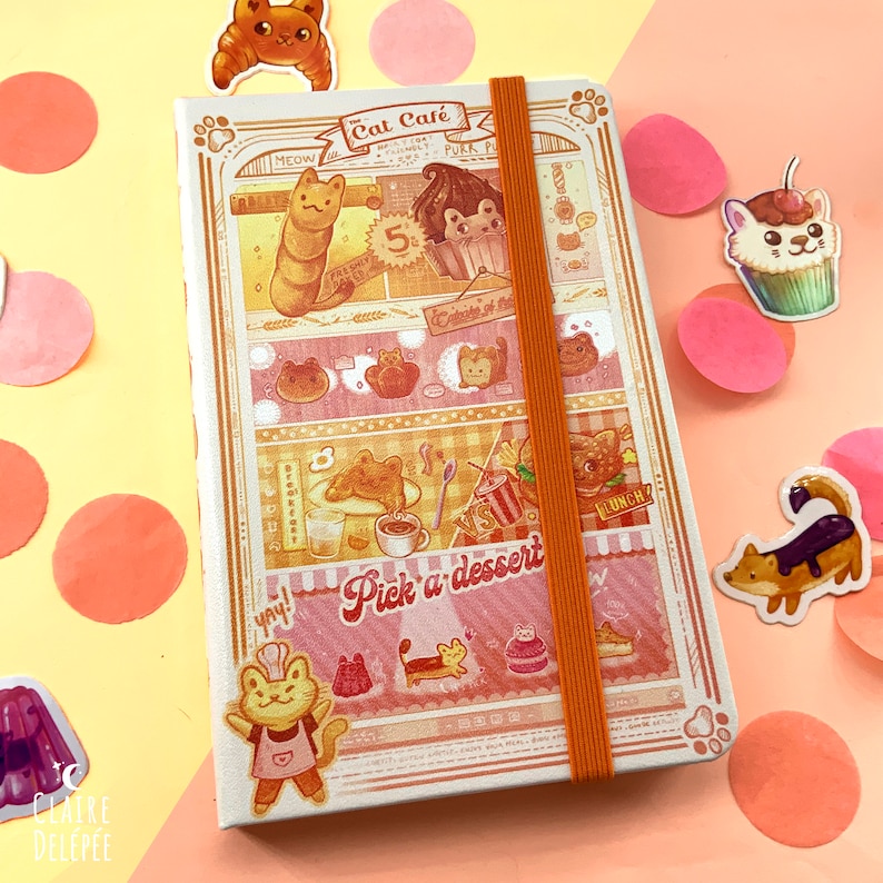 Puede incluir: Un cuaderno ilustrado rosa y naranja con tem&aacute;tica de cafeter&iacute;a de gatos. La portada presenta un gato con un gorro de chef y sosteniendo un batidor, con el texto "Cat Cafe" encima. Hay otras ilustraciones de gatos y postres en la portada.