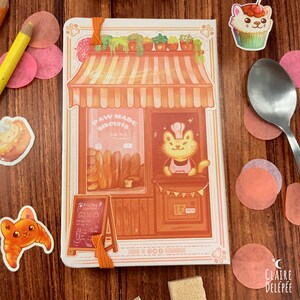 Puede incluir: Una libreta blanca con una ilustraci&oacute;n de una panader&iacute;a de gatos. La panader&iacute;a tiene un letrero que dice "Paw Made Biscuits". El gato lleva un gorro de chef y un delantal. La libreta tiene una cinta naranja.