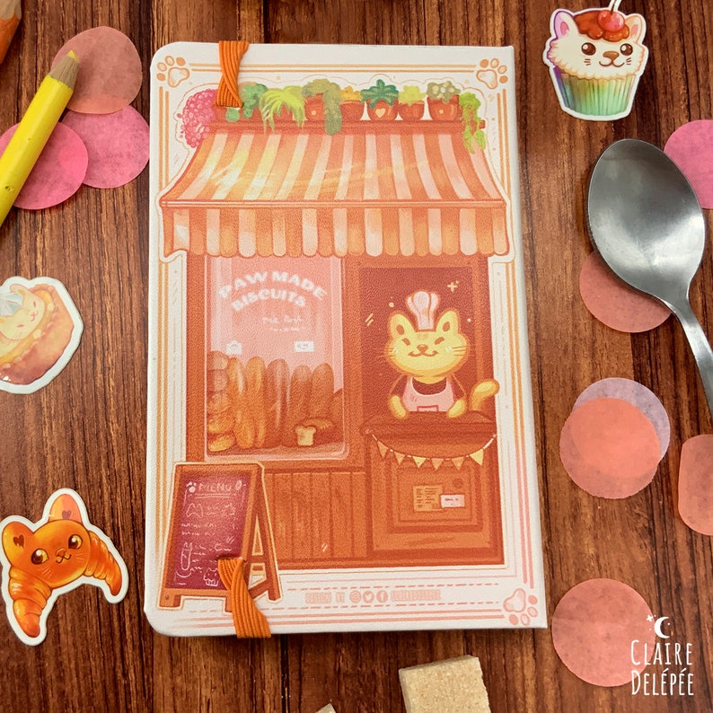 Puede incluir: Una libreta blanca con una ilustraci&oacute;n de una panader&iacute;a de gatos. La panader&iacute;a tiene un letrero que dice "Paw Made Biscuits". El gato lleva un gorro de chef y un delantal. La libreta tiene una cinta naranja.