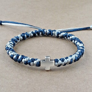 May include: A braided cord bracelet featuring a central silver-toned metal cross charm. The bracelet is constructed from alternating dark navy blue and light grey nylon threads, with two loose cord ends extending from the adjustable sliding knot closure.