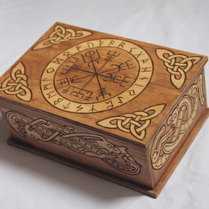 Peut inclure: Une boîte rectangulaire en bois avec une finition brun foncé et une pyrogravure complexe. Le couvercle présente un symbole Vegvisir circulaire entouré de runes, flanqué de nœuds celtiques aux coins. Les panneaux latéraux montrent des motifs de serpents.