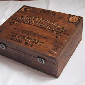 Könnte beinhalten: Eine rechteckige Holzbox mit dunkelbrauner Oberfläche, die auf dem Deckel ein eingebranntes Ouija-Brett-Design aufweist. Der Deckel zeigt das Alphabet, die Zahlen 1-9, 0, die Wörter YES, NO, GOOD BYE sowie Sonnen- und Mondmotive und Metallverschlüsse.