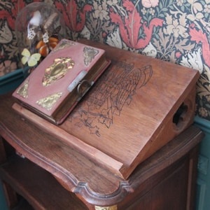 May include: A wooden desk with a book stand, featuring a book with a pink cover and gold accents. The desk has a carved design and is set against a floral wallpaper background. A glass dome with decorative elements sits on top.