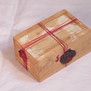 Op de afbeelding: Een rechthoekige houten doos met een lichtbruine afwerking, omwikkeld met dun rood touw dat over de bovenkant kruist. De doos is voorzien van twee donkerrode waszegels aan de zijkant en een vage cirkelvormige stempel op de houten voorzijde.