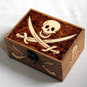 Puede incluir: Caja de madera con temática pirata, con una calavera y huesos cruzados en la tapa. La caja tiene un acabado marrón oscuro y un cierre de metal. La calavera y las espadas son de color claro.