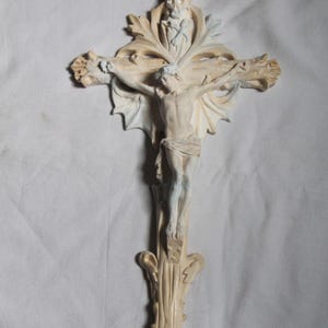 Op de afbeelding: Een crèmekleurig kruisbeeld met een gedetailleerde gravure van Jezus Christus aan het kruis. Het kruis heeft sierlijke, bladachtige ontwerpen en een decoratieve basis. De figuur van Christus is beschilderd met subtiele blauwe en witte accenten.
