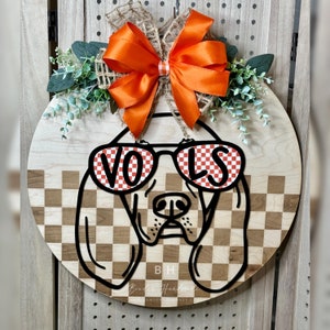 May include: A round wooden door hanger with a black and white checkered pattern. A black silhouette of a dog wearing sunglasses with the word "VOLS" in red and white checkered letters is in the center of the door hanger. An orange bow with a burlap center is on top of the door hanger.