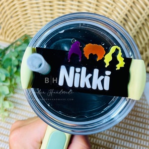 May include: A black and white personalized tumbler with a green lid and straw. The tumbler has a sticker with three colorful witch silhouettes and the name "Nikki" on it.