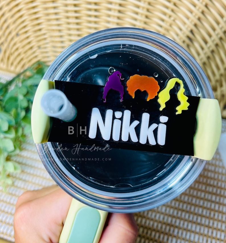 May include: A black and white personalized tumbler with a green lid and straw. The tumbler has a sticker with three colorful witch silhouettes and the name "Nikki" on it.