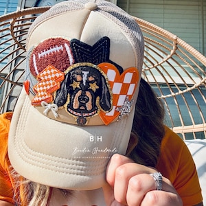 May include: A beige trucker hat with a mesh back. The hat has a variety of patches on the front, including a football, a hound dog, a heart with a checkered pattern, and a bow. The hat also has the text "Bowden Handmade" and "B.H." on the front.