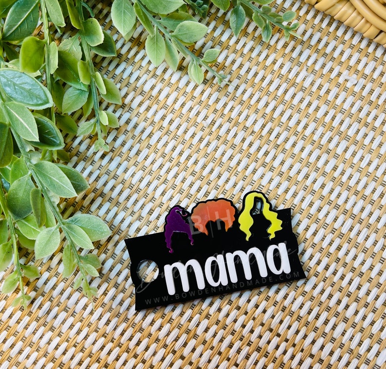 May include: Black acrylic keychain with three silhouettes of women with colorful hair and the word "mama" in white text. The text "www.bowdenhandmade.com" is printed below the word "mama".