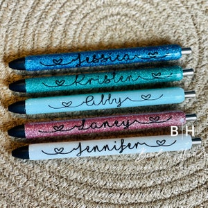 Puede incluir: Cinco bol&iacute;grafos de purpurina personalizados con los nombres "Jessica", "Kristen", "Abby", "Janey" y "Jennifer" escritos en tinta negra. Cada bol&iacute;grafo tiene un peque&ntilde;o s&iacute;mbolo de coraz&oacute;n al lado del nombre.