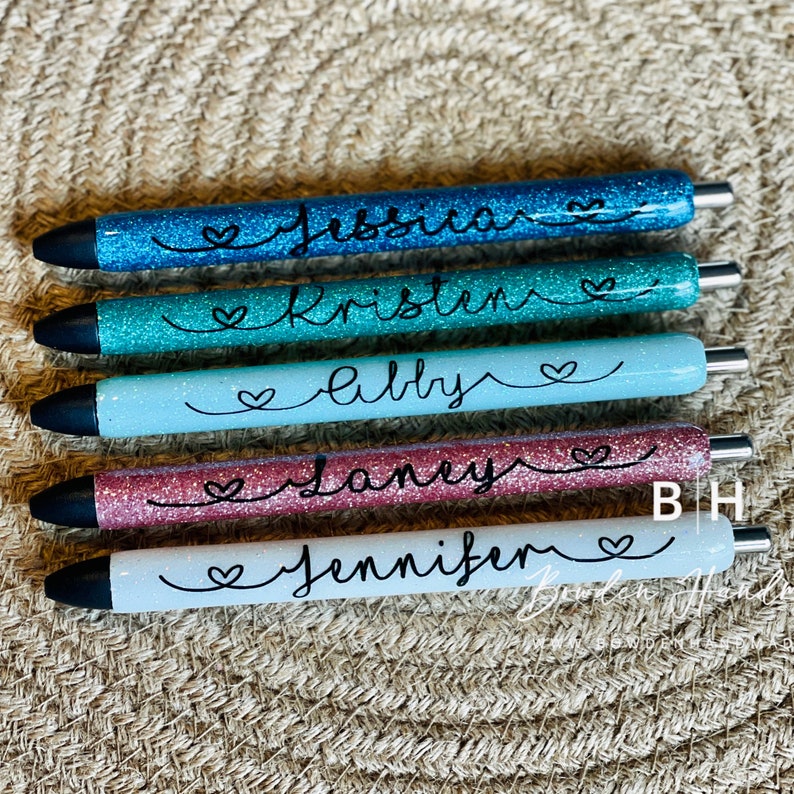Puede incluir: Cinco bol&iacute;grafos de purpurina personalizados con los nombres "Jessica", "Kristen", "Abby", "Janey" y "Jennifer" escritos en tinta negra. Cada bol&iacute;grafo tiene un peque&ntilde;o s&iacute;mbolo de coraz&oacute;n al lado del nombre.