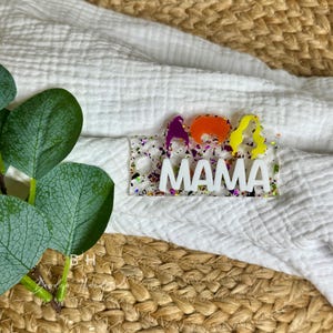 May include: A white acrylic keychain with colorful glitter embedded in the background. The keychain says "MAMA" in white letters and has three colorful cartoon figures on the left side.