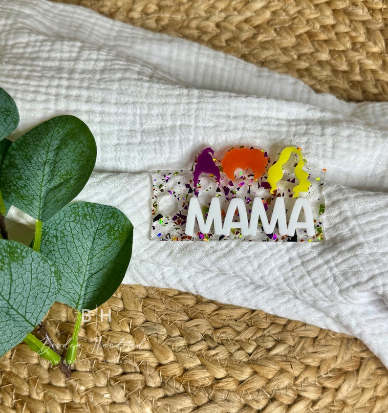 May include: A white acrylic keychain with colorful glitter embedded in the background. The keychain says "MAMA" in white letters and has three colorful cartoon figures on the left side.