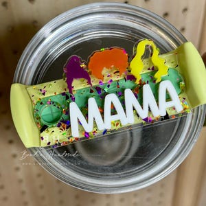 May include: A clear glass tumbler with a green, yellow, and purple glitter lid. The lid has a white vinyl sticker with the word "MAMA" in black letters. The lid also has a clear plastic sticker with a silhouette of four women in different colors.