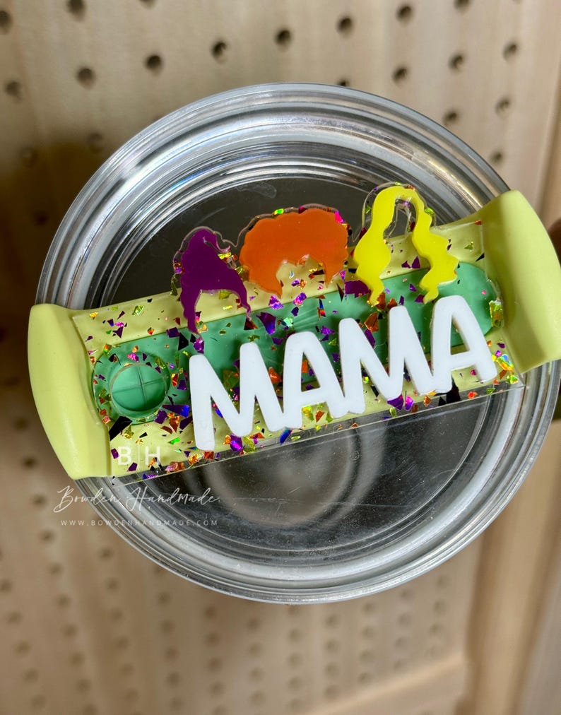May include: A clear glass tumbler with a green, yellow, and purple glitter lid. The lid has a white vinyl sticker with the word "MAMA" in black letters. The lid also has a clear plastic sticker with a silhouette of four women in different colors.
