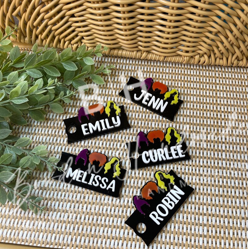 May include: Five black acrylic keychains with colorful cartoon images and white text. The names on the keychains are Jenn, Emily, Curlee, Melissa, and Robin.