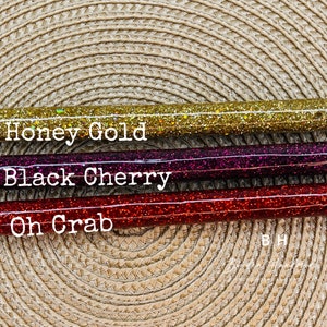 Puede incluir: Tres bol&iacute;grafos de purpurina con tapas negras y detalles plateados. Los bol&iacute;grafos est&aacute;n etiquetados como "Honey Gold", "Black Cherry" y "Oh Crab".